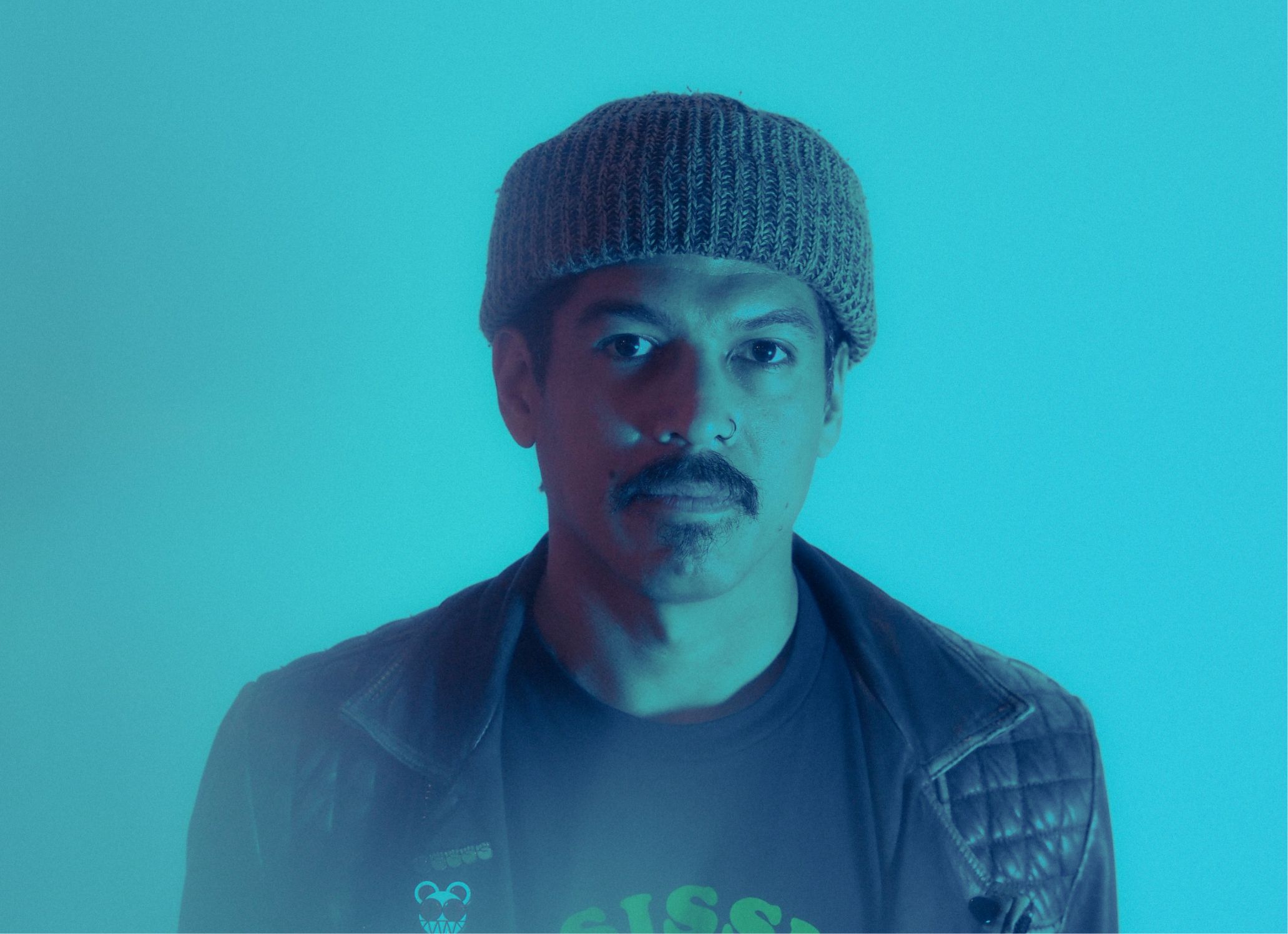 A man with a mustache wearing a beanie and leather jacket against a blue background.