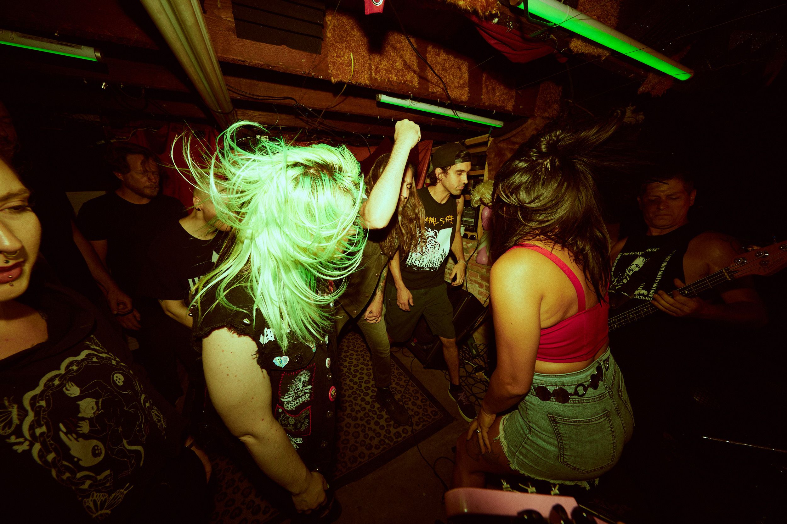 A group of people at a lively concert or music event, with vibrant lighting and a person in the foreground flipping their light-colored hair.