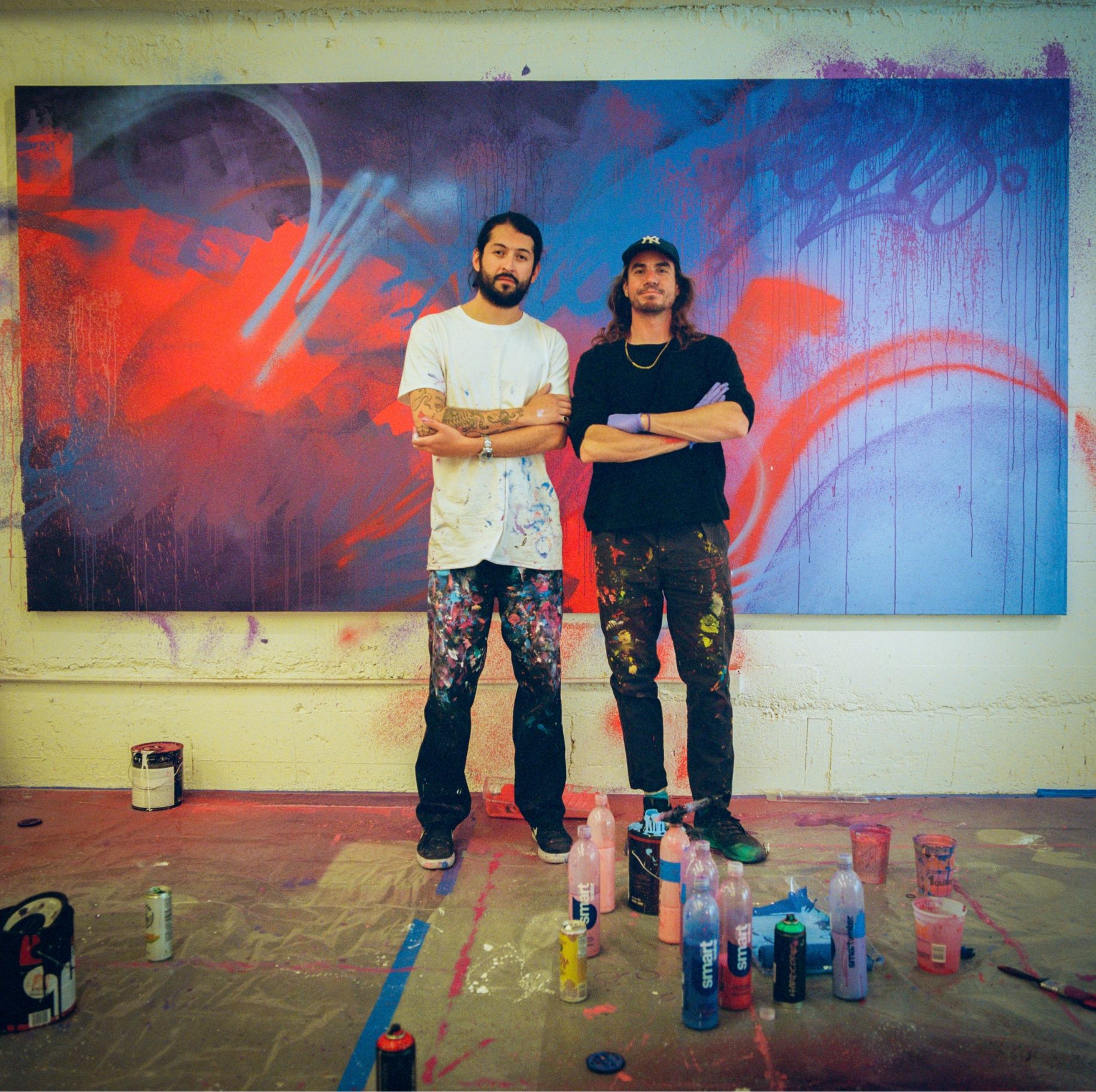 Madsteez and Its a Living stand in front of finished large painting. Paint supplies and drop cloths cover floor