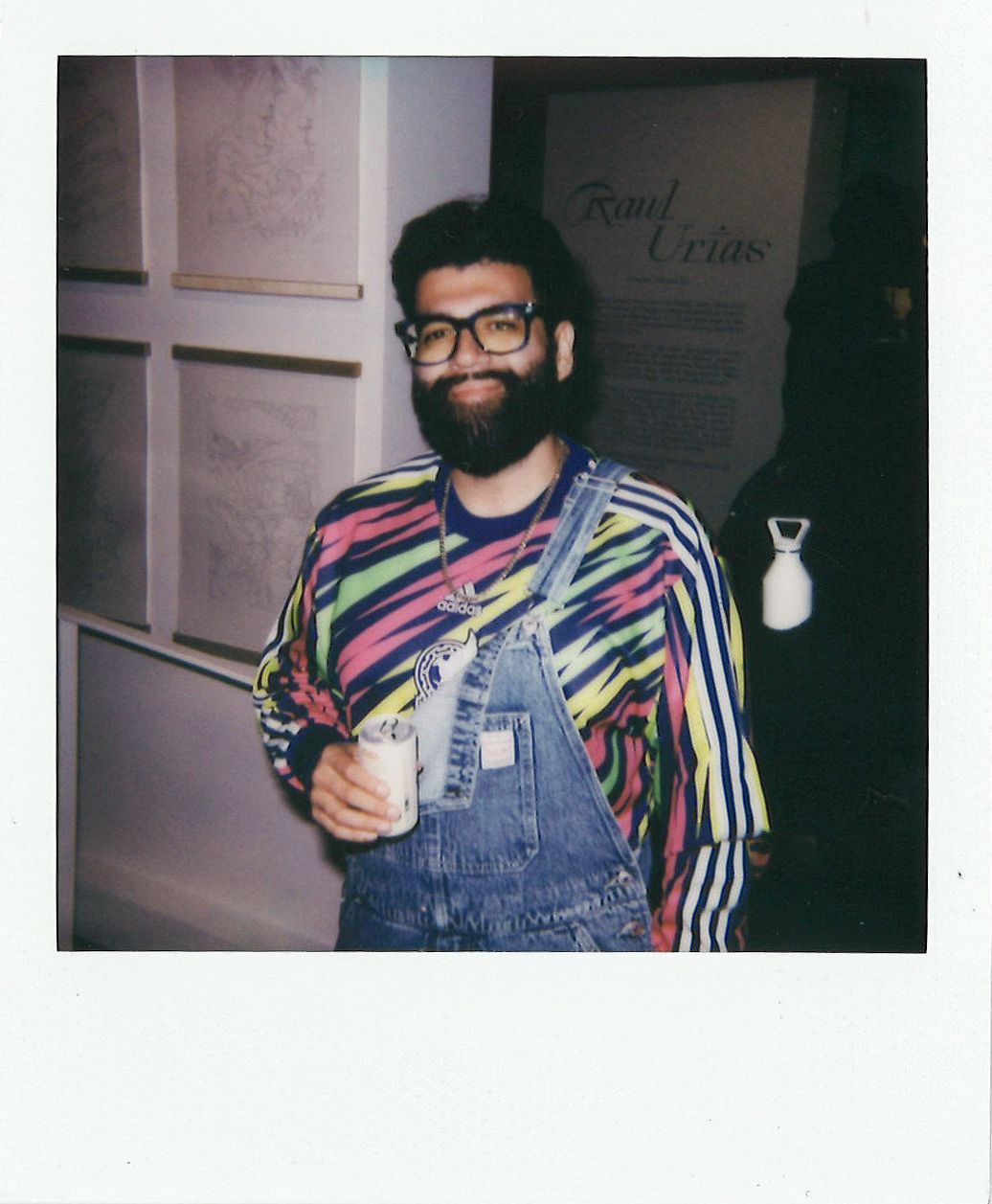 A person with glasses and a beard wearing a colorful shirt and overalls holds a drink at an art exhibit.