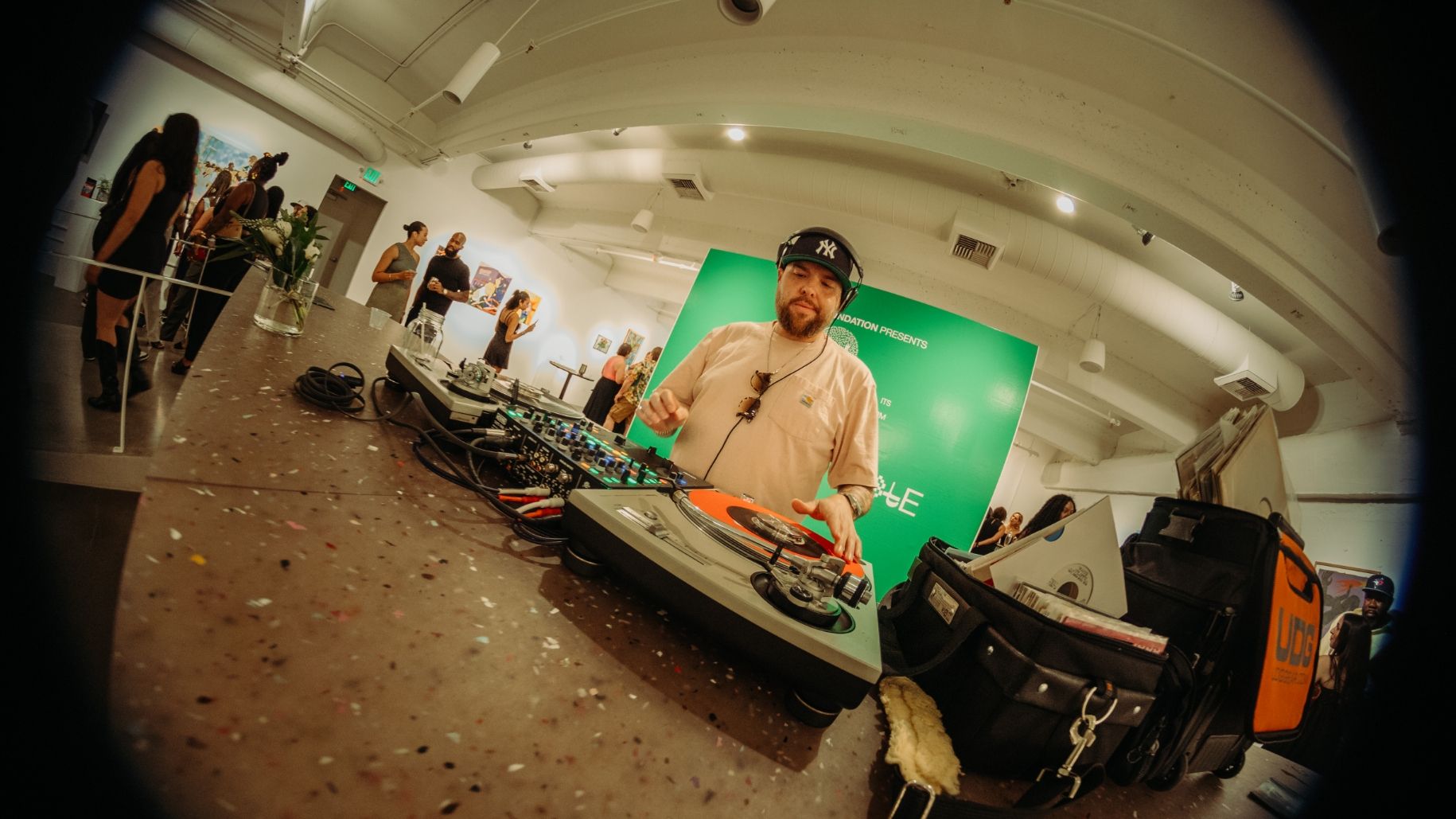 A man in a beige shirt and black cap is DJing on turntables in a modern art gallery with a green wall in the background. Equipment cases are visible around him. Visitors are seen in the gallery.