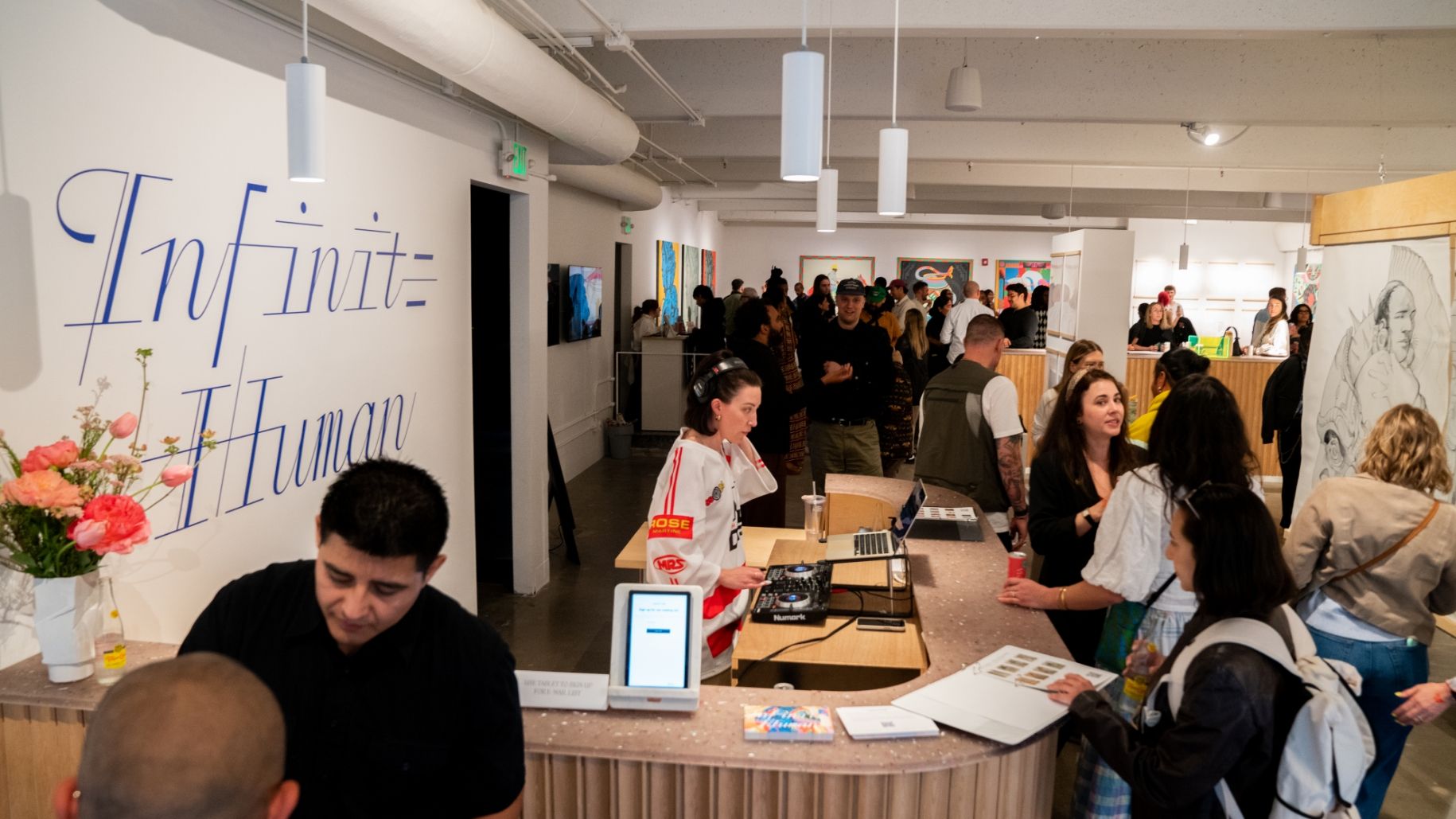 People gather in a modern art gallery space with artwork displayed on the walls and a counter with a DJ setup and flowers. The phrase "Infinite Human" is written in large letters on the wall.