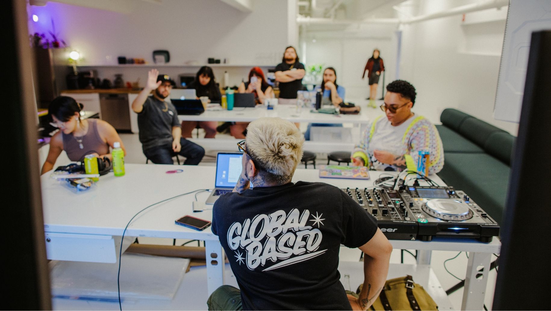 A group of people are gathered in a well-lit, modern room. One person with a "GLOBAL BASED" shirt is seated at a table with DJ equipment, while others sit near laptops and raise hands.