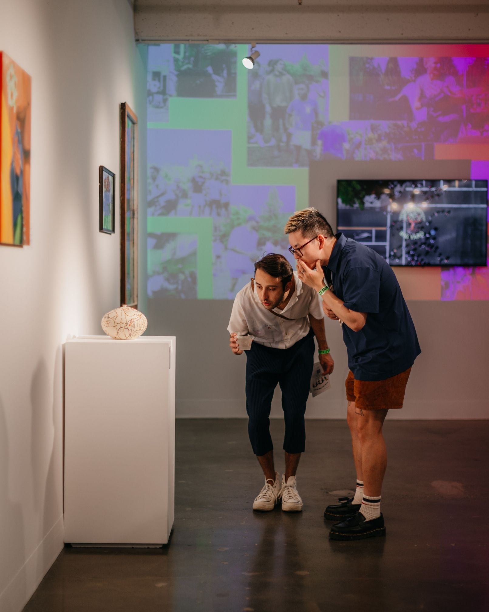 Two people closely examining an object displayed on a pedestal in a contemporary art gallery with various artworks on the walls and a projected collage in the background.