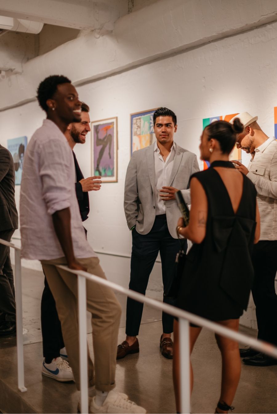 A group of people casually dressed stand and converse in an art gallery, with colorful artworks displayed on white walls in the background.