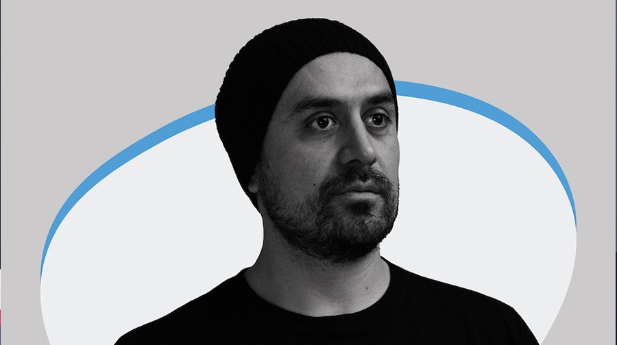 A grayscale portrait of a man wearing a beanie and a black shirt, with a neutral expression. A blue and white abstract shape is in the background.