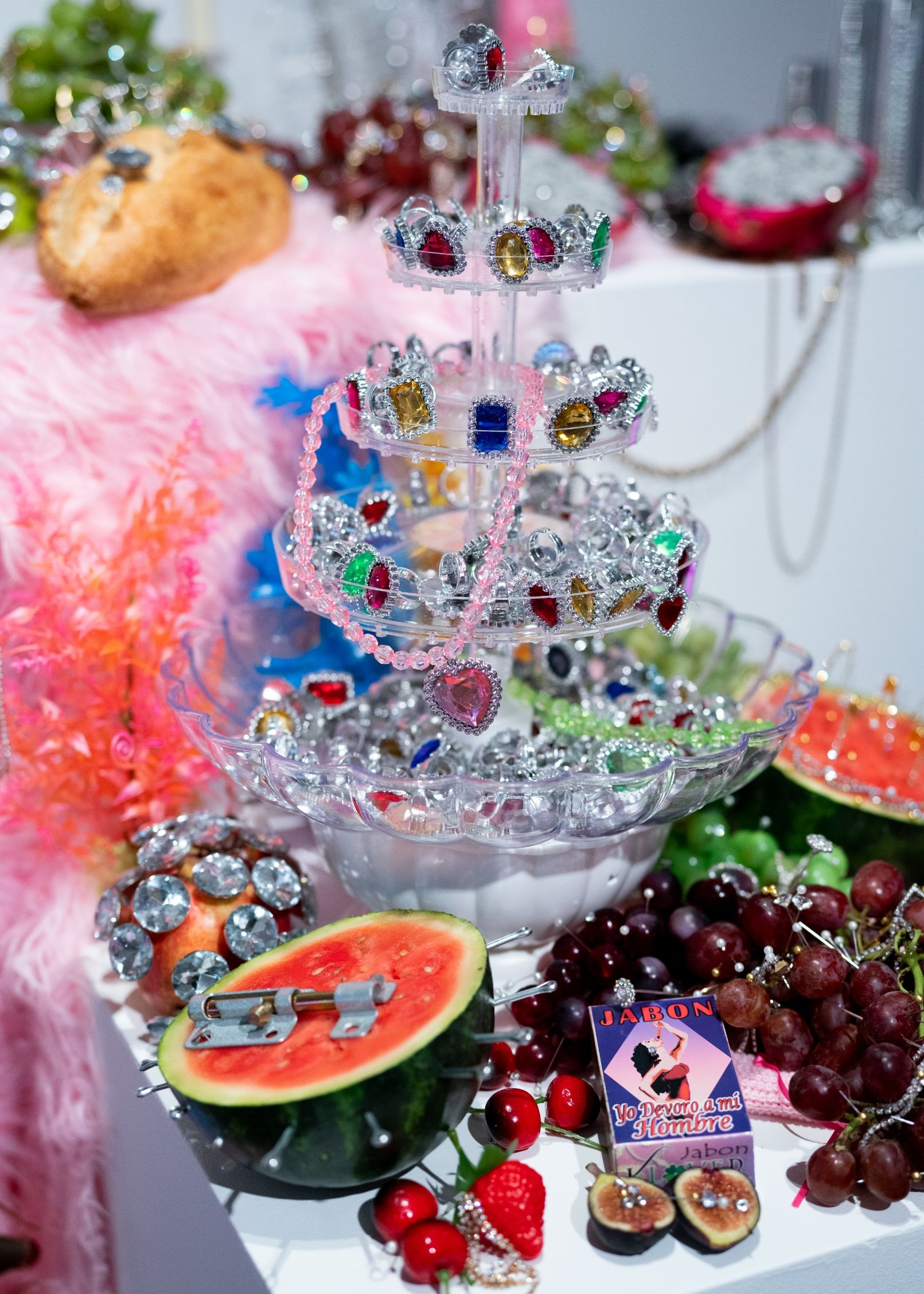 A decorative display with a tiered stand of colorful rings, surrounded by grapes, cherries, half a watermelon, a feather boa, and a small box labeled "Baron.