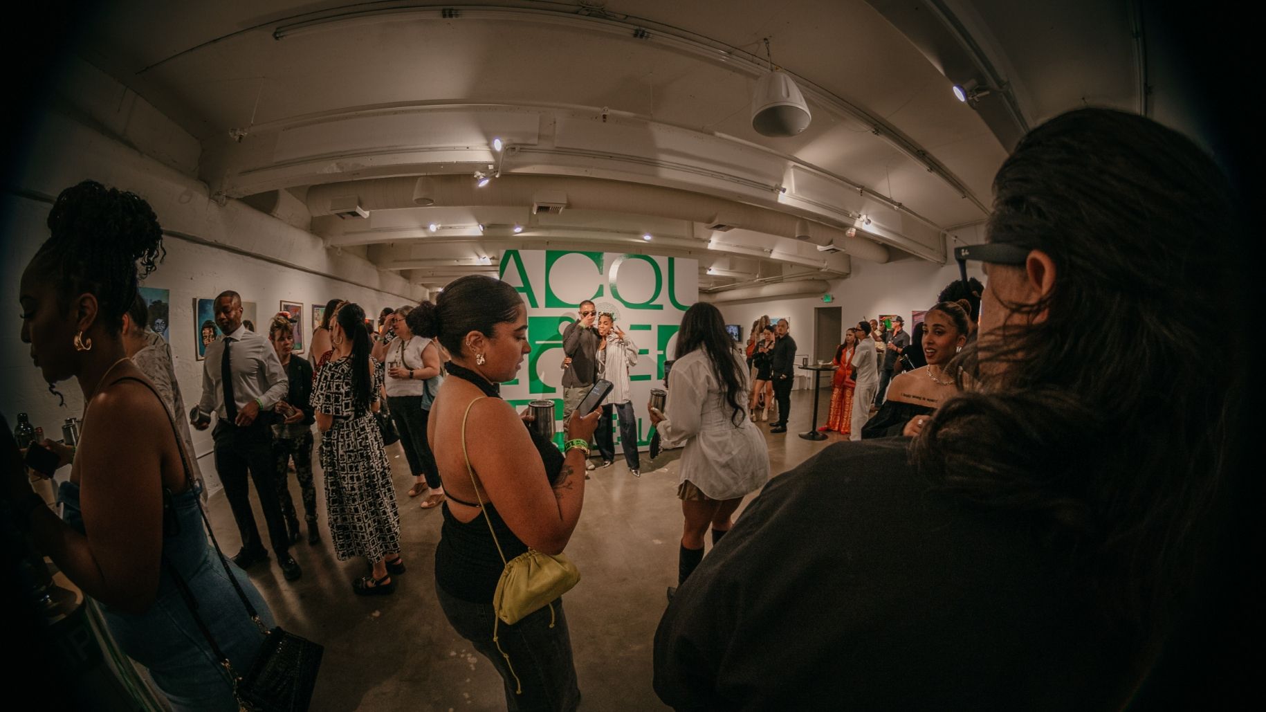 A group of people socializes in an art gallery with "ACQUISITIONS" written on the wall in the background.