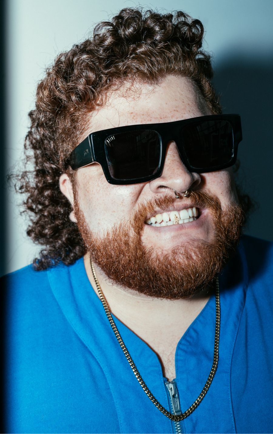 Close-up of a smiling man with curly hair and beard, wearing large black sunglasses and a blue shirt.