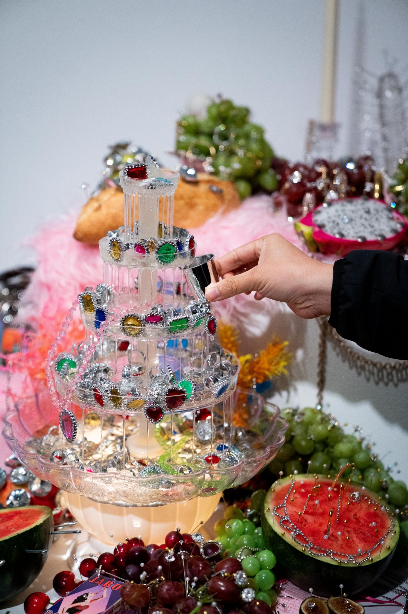 A hand adjusts crystals on a decorative glass tiered display, surrounded by grapes, watermelon slices, and other fruits.