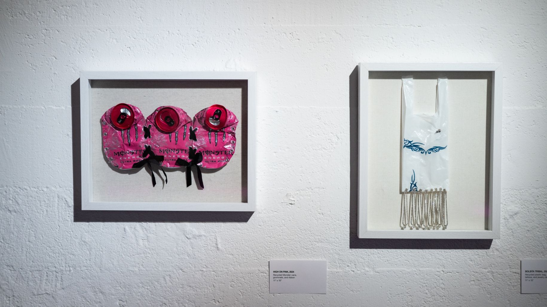 Two framed artworks on a white wall; the left piece features pink elements with text and ribbons, the right piece features a white item with blue design and hanging chains.