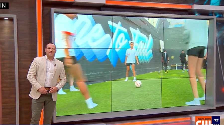 A news anchor stands beside a large screen displaying people playing soccer on a green field in front of a mural.