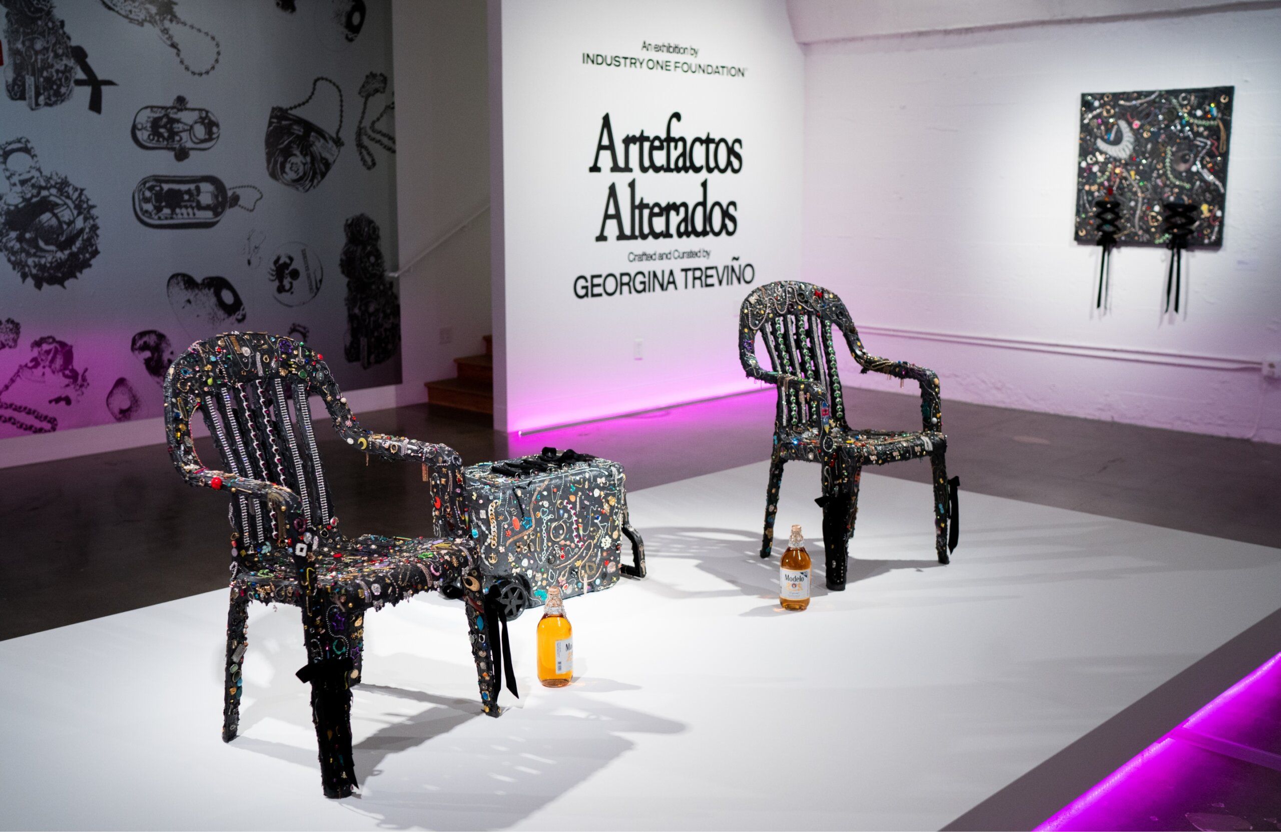 Two intricately beaded chairs with various patterns are displayed on a white platform in a gallery setting. There are two bottles on the floor, and a similar artwork is displayed on a wall.