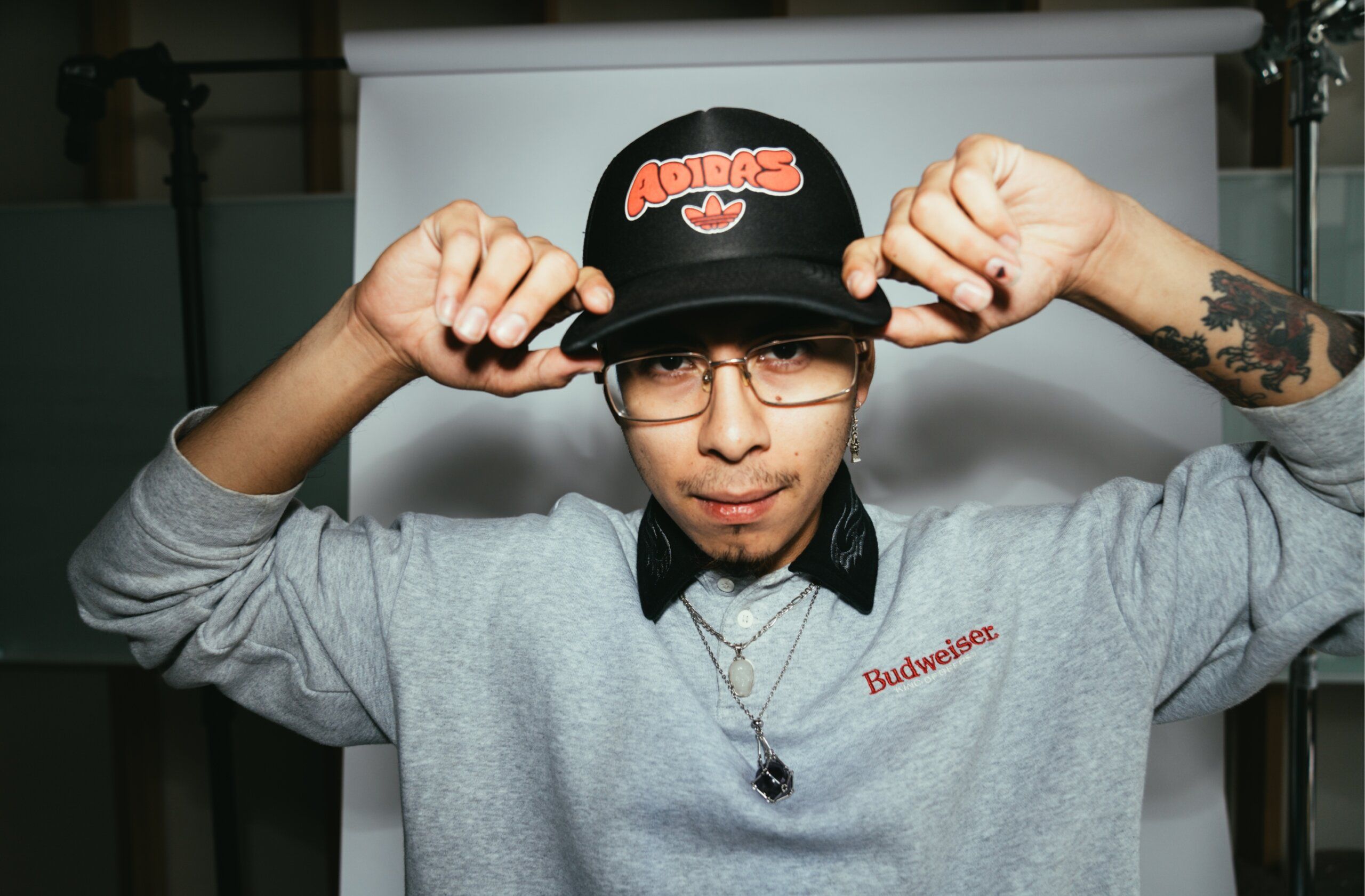 A young man adjusts his black Adidas cap, wearing glasses, a bow tie, and a grey Budweiser sweatshirt, standing in front of a white backdrop.