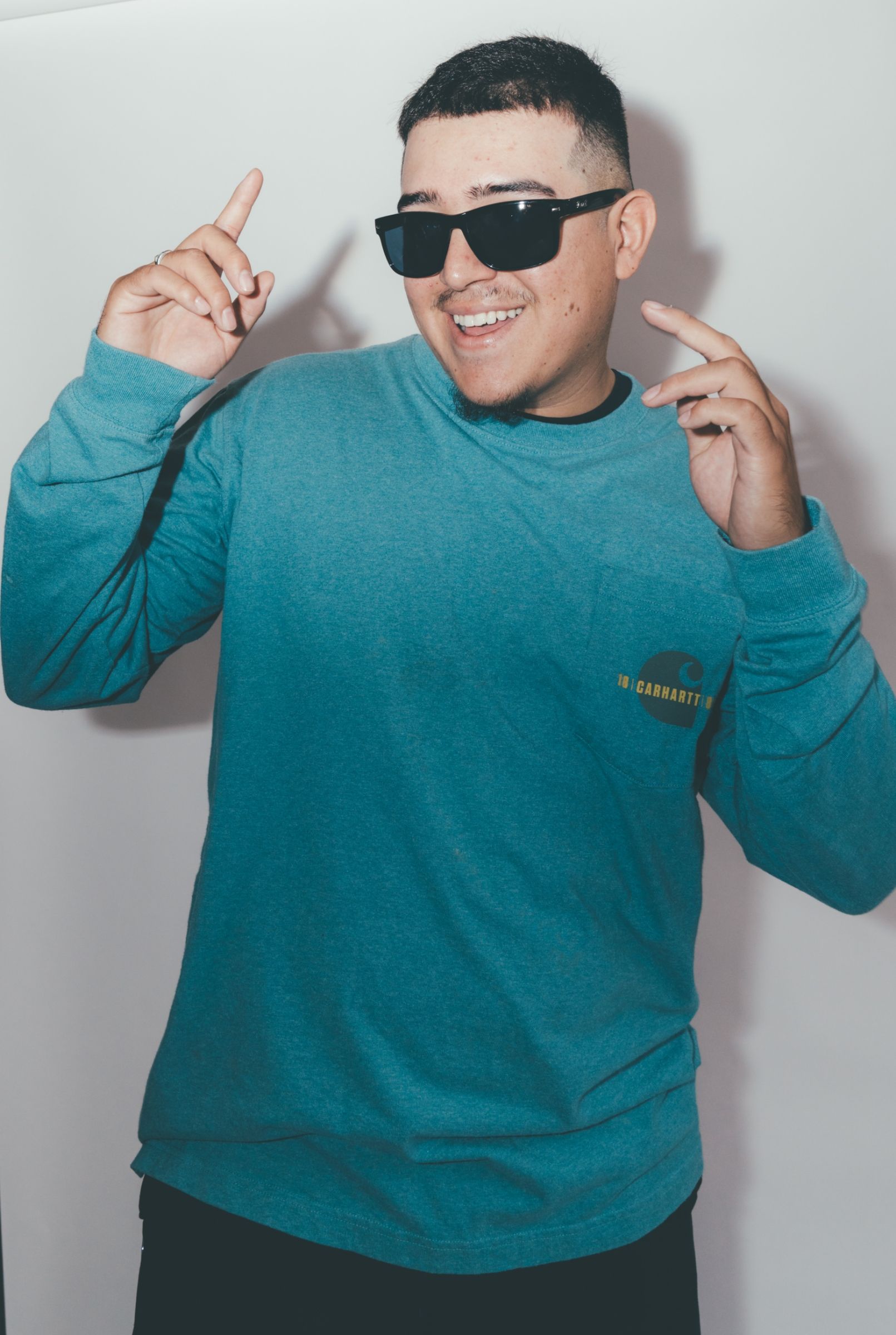 A smiling man wearing sunglasses and a blue Carhartt sweatshirt, dancing and pointing upwards with his fingers.