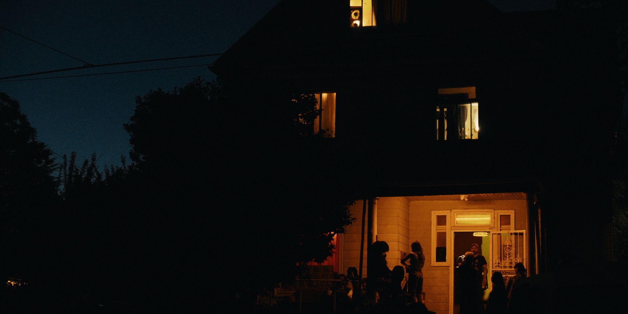 A house at night with warm light glowing from the windows and porch, people visible inside and near the entrance.