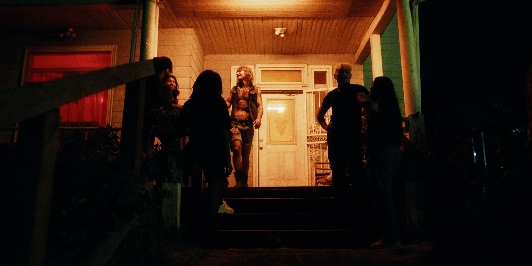 A group of people stand on the steps of a porch at night, illuminated by warm light from the house.