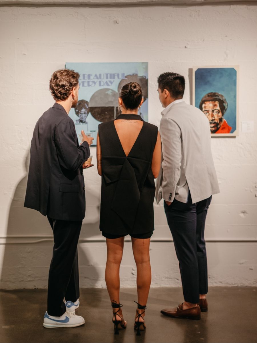 Three individuals are standing in a gallery, viewing artwork on a white wall. The artwork appears to be colorful and portrait-oriented. The setting is well-lit with a polished concrete floor.