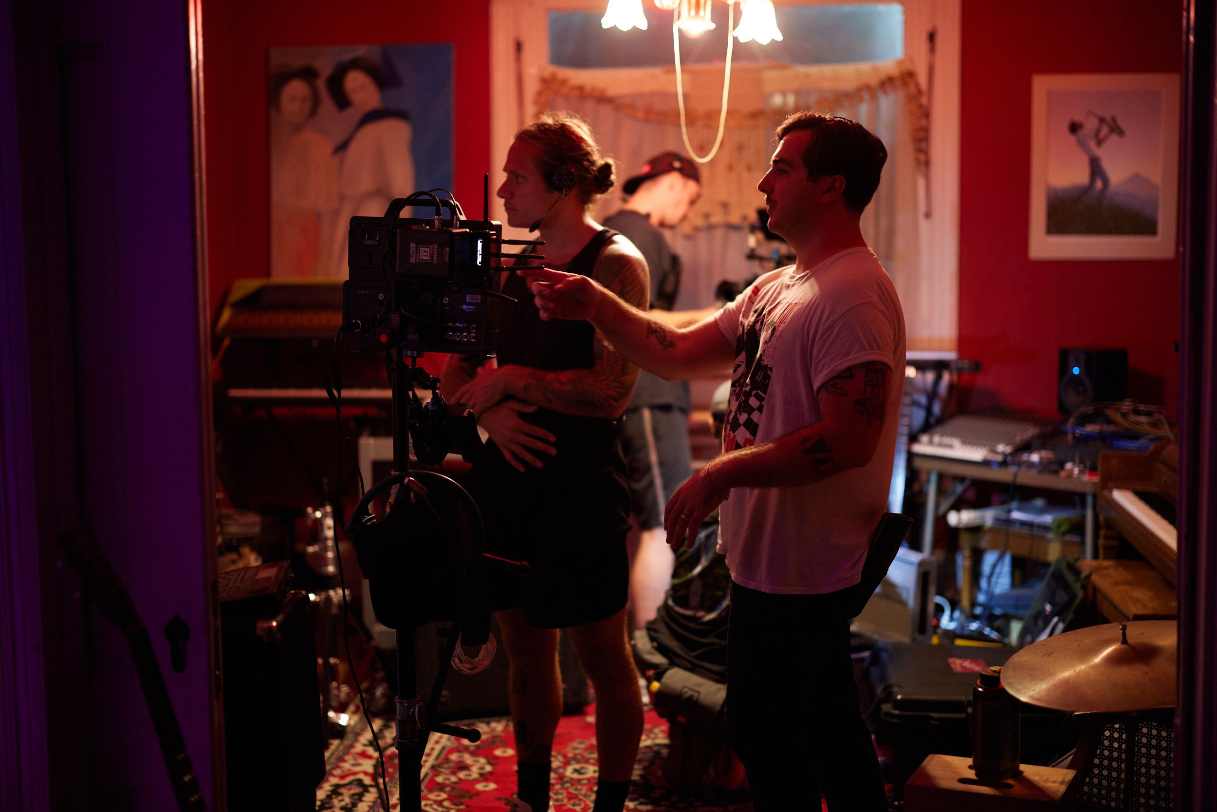 Two people work with camera equipment in a dimly lit room with musical instruments and artwork on the walls.