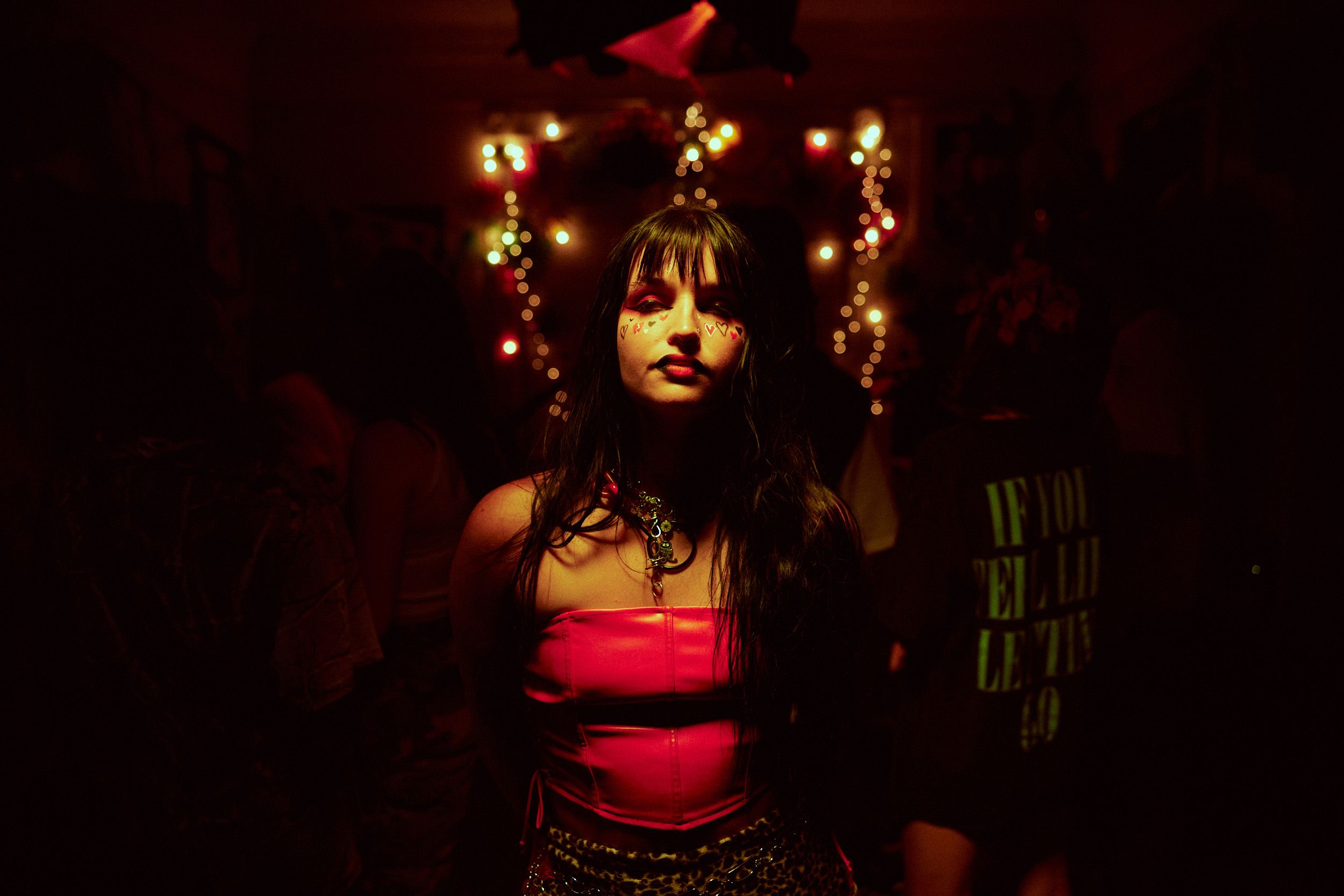 A woman in a red top stands under dim lighting with multicolored lights in the background at a party.