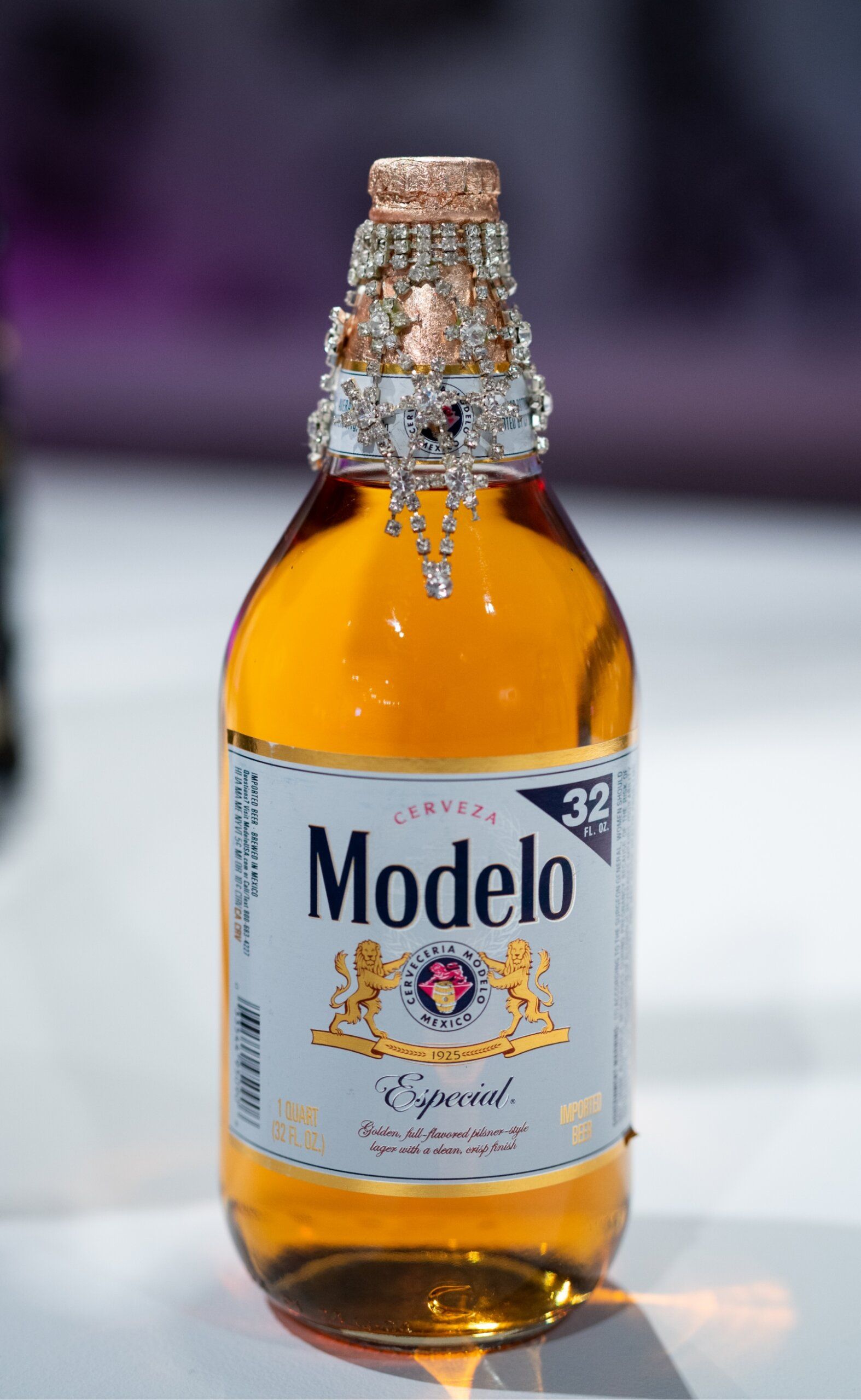 A 32 fl. oz. bottle of Modelo Especial beer with a decorative beaded cap on a blurred background.