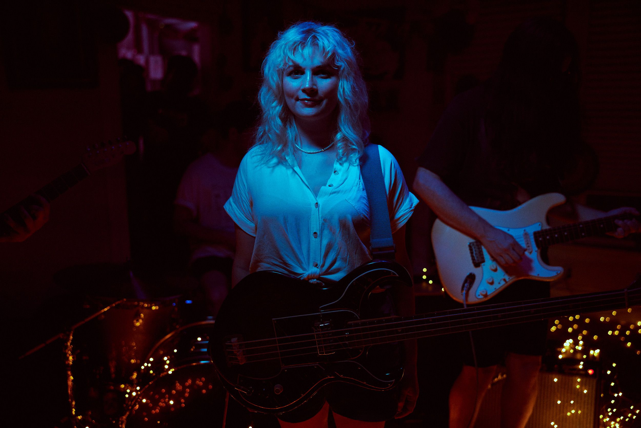 A person with blonde hair plays a bass guitar in a dimly lit room, surrounded by other musicians with instruments.