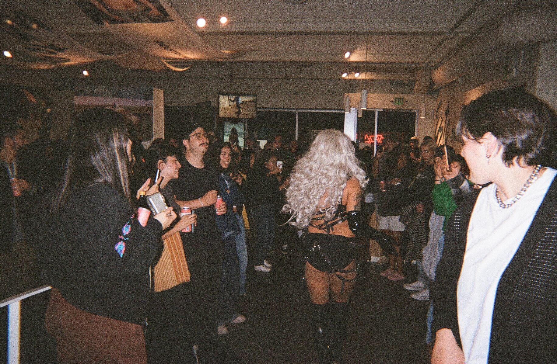 drag performer in light blond wig and walks by a group of people at art gallery while doing a drag performance
