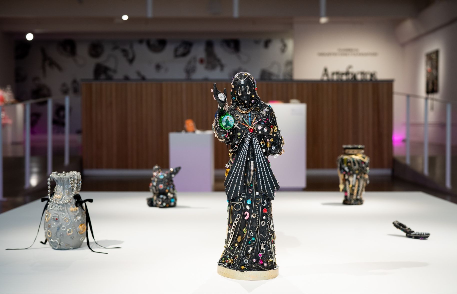 Art exhibit featuring sculptures adorned with various embellishments, displayed on a white platform in a gallery setting with a wood-paneled background.