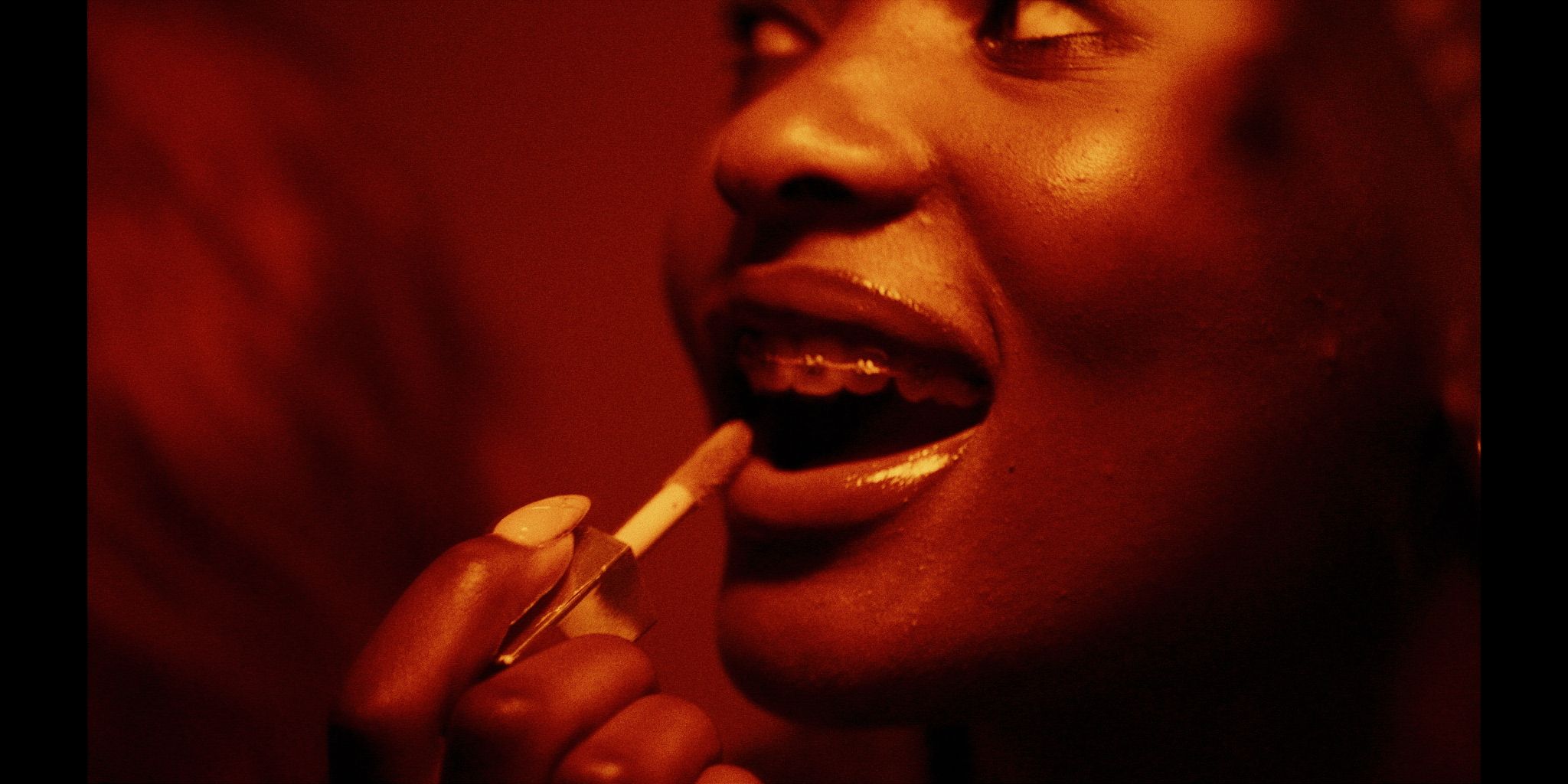 Close-up of a person applying lipstick, highlighting braces on their teeth. The image is lit in warm, orange tones.