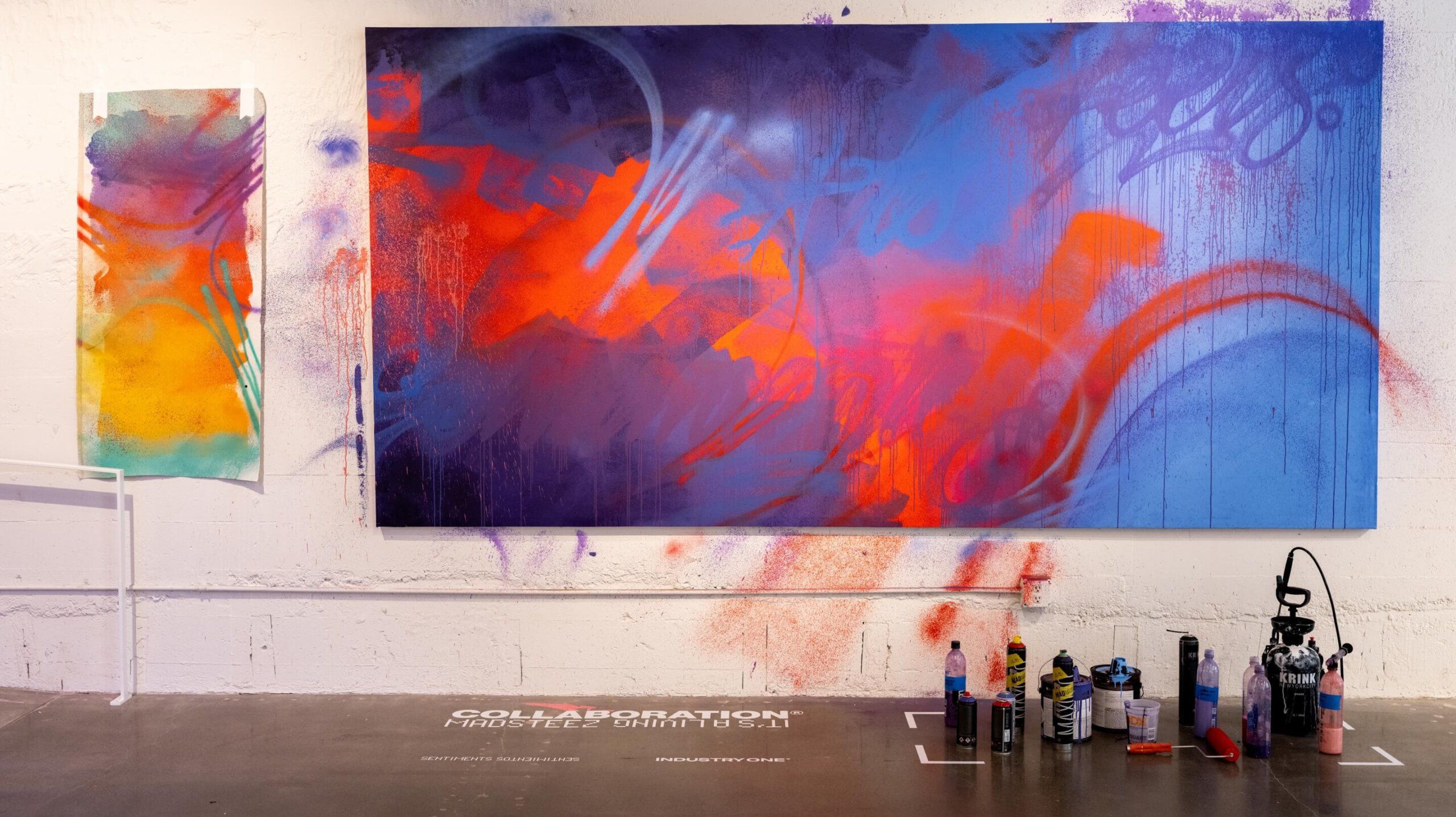 A color test spray painted sheet of paper taped next to a large colorful painting. Painting supplies are on the floor in front of painting as part of the exhibit