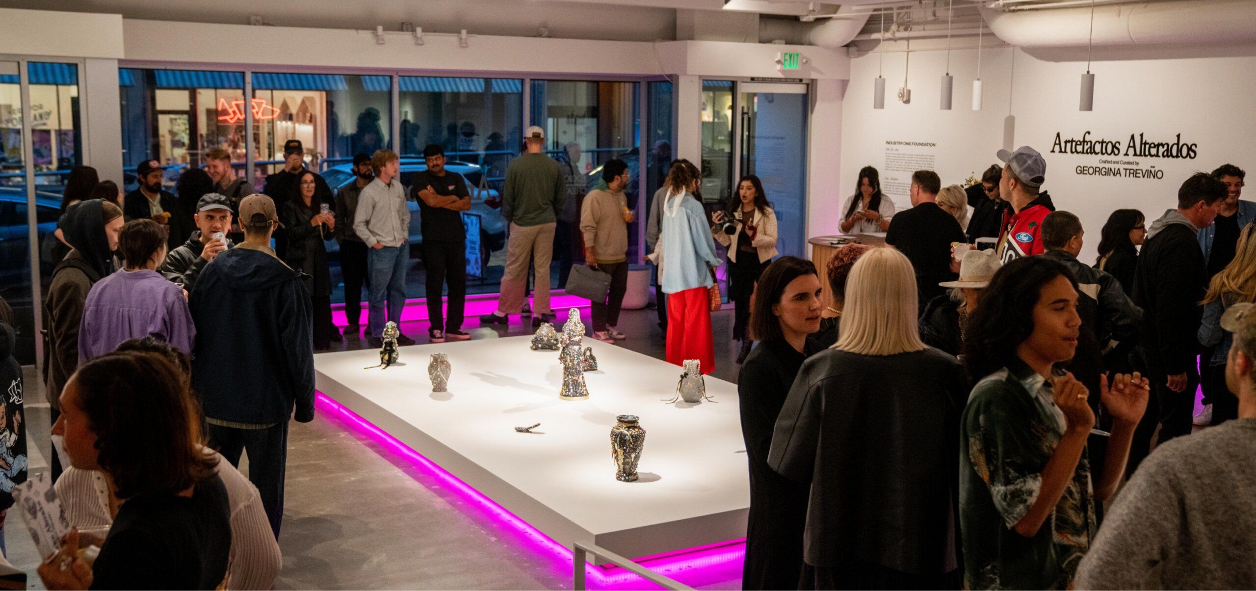 People attend an art exhibition in a gallery with sculptures displayed on a lit platform. The walls have text referring to "Artefactos Alterados" by Georgina Treviño.