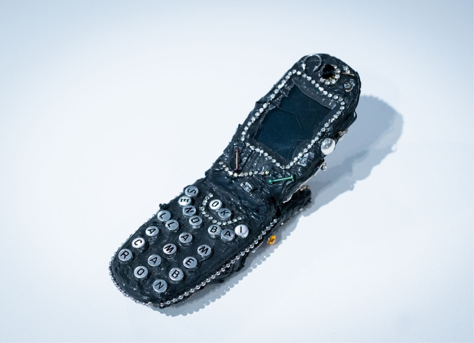 A handmade model of a flip phone, decorated with beads and wires, is displayed on a plain surface.