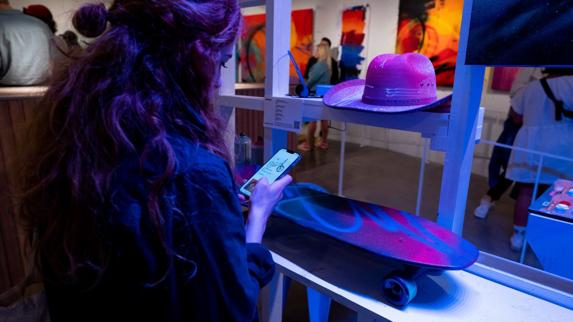 Women looks up artwork prices and info on her phone while in front of spray painted skateboard and spray painted cowboy hat
