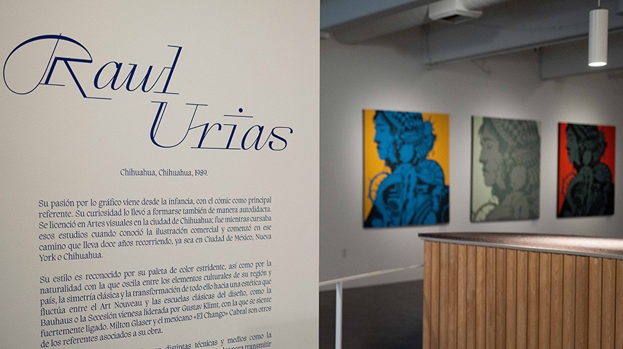 Exhibit description about Raul Urias next to three of his artworks featuring stylized portraits hung on a gallery wall. The room has a contemporary design and the artworks are prominently displayed.