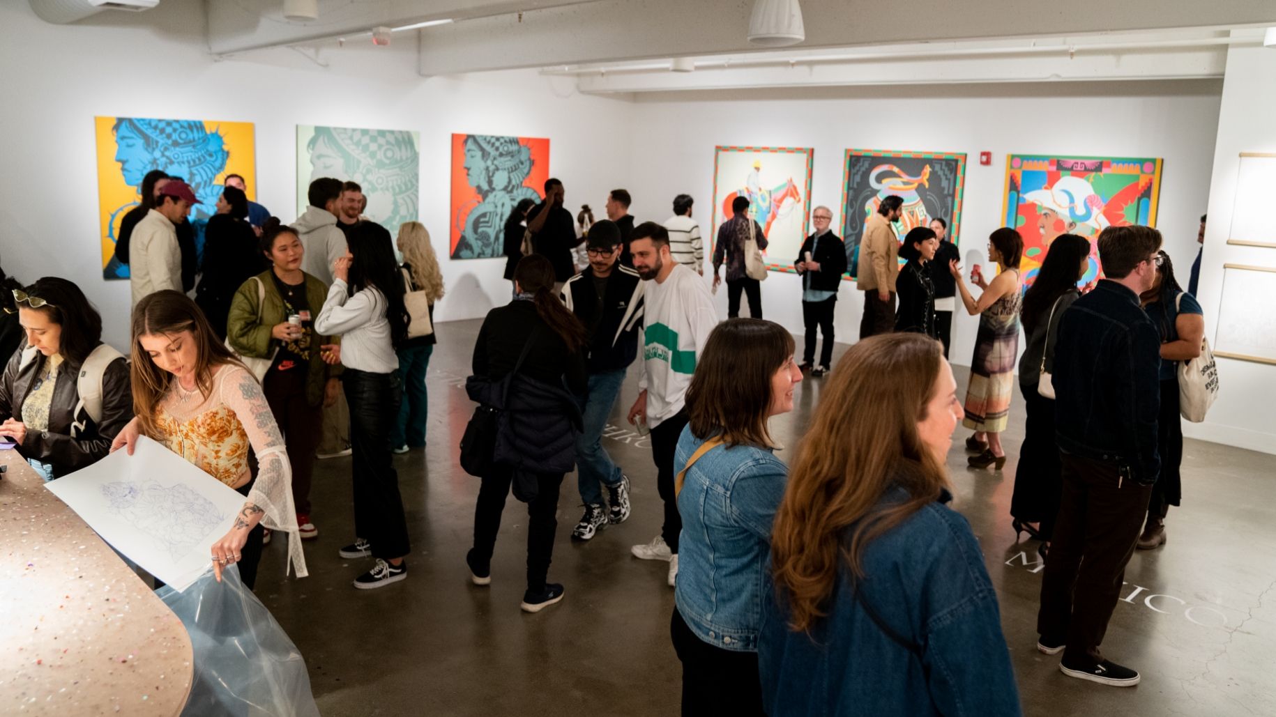 A crowd of people in an art gallery observe and discuss colorful artworks displayed on the walls. Some individuals hold papers, and the gallery space is brightly lit.