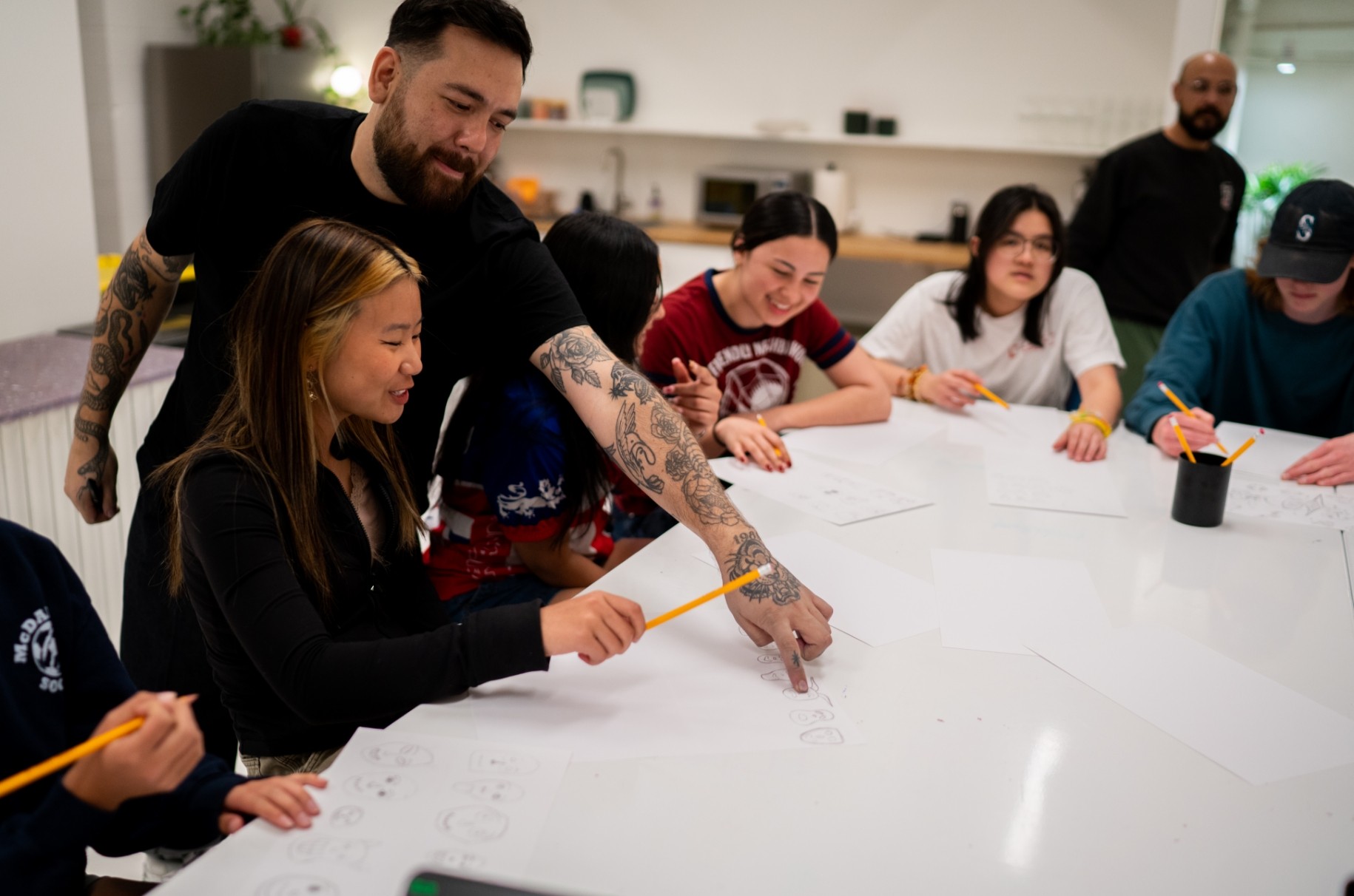 Adults gathered around a table in a workshop, drawing on paper with pencils and markers, interacting and smiling.