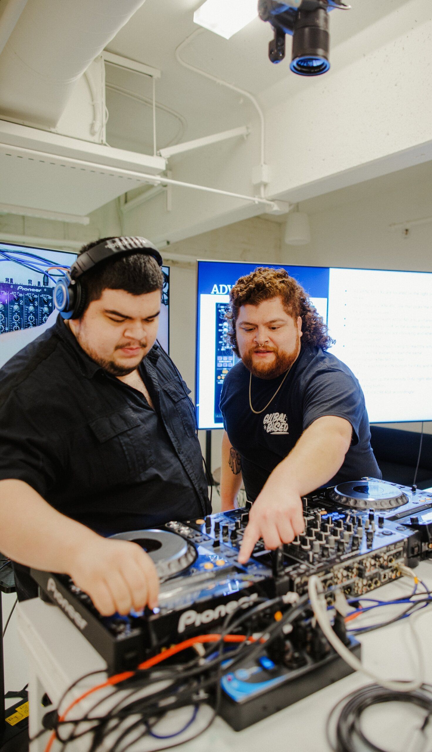Two men operate DJ equipment, with one wearing headphones and the other pointing at a control panel. They are in an indoor setting with a screen displaying text in the background.
