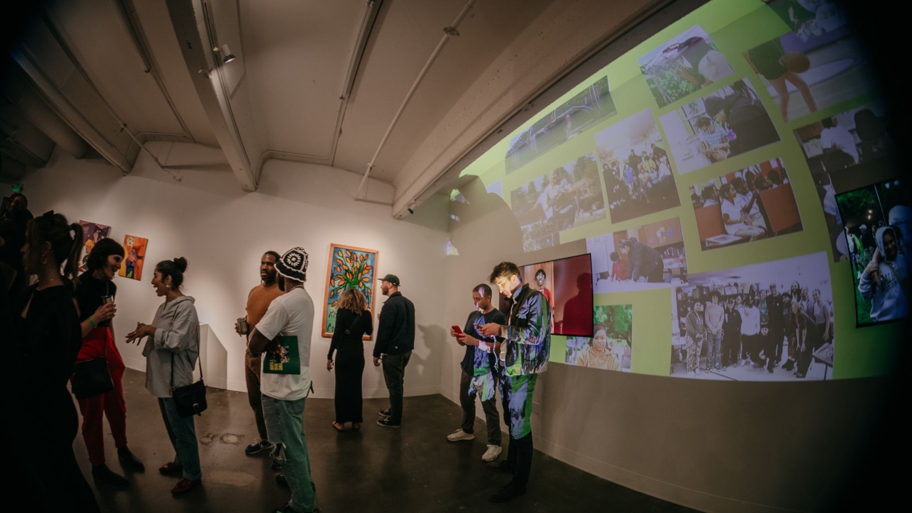 A group of people standing and conversing in an art gallery, with various paintings on the wall and a projection of photo collages on a large screen.