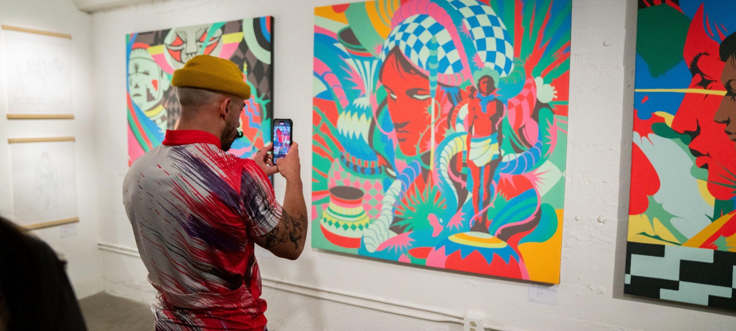 A person in a colorful shirt and yellow hat takes a photo of vibrant, abstract art displayed on a wall in an art gallery.