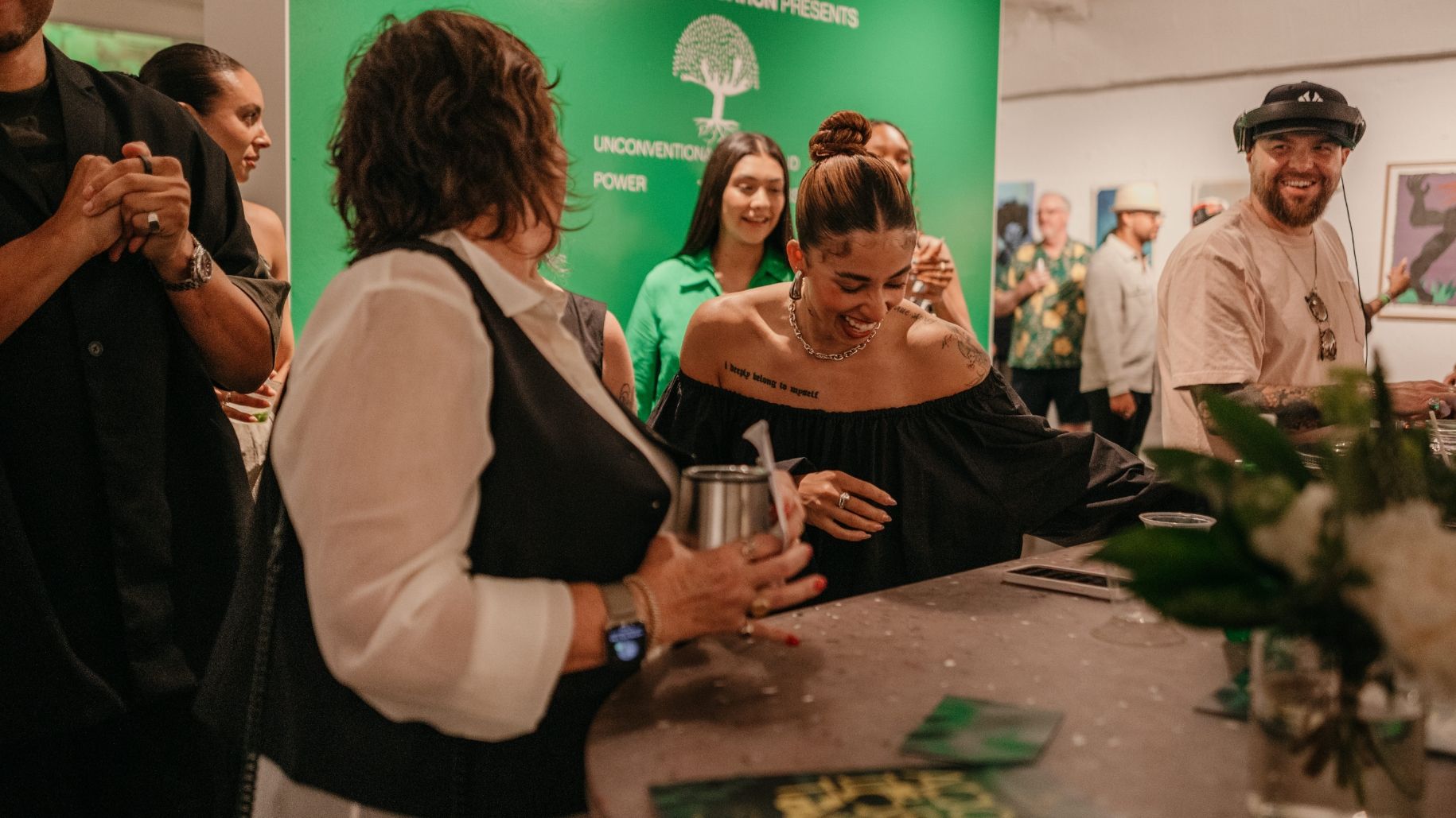 A group of people, some smiling and others engaged in conversation, stand around a table in a room with green walls and art displays. Foreground shows a woman with a tattoo on her shoulder holding a drink.