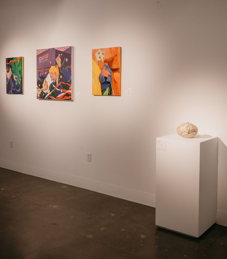 A gallery space with four colorful abstract paintings on the wall and a sculptural piece displayed on a white pedestal. The room is well-lit, showcasing the artwork prominently.