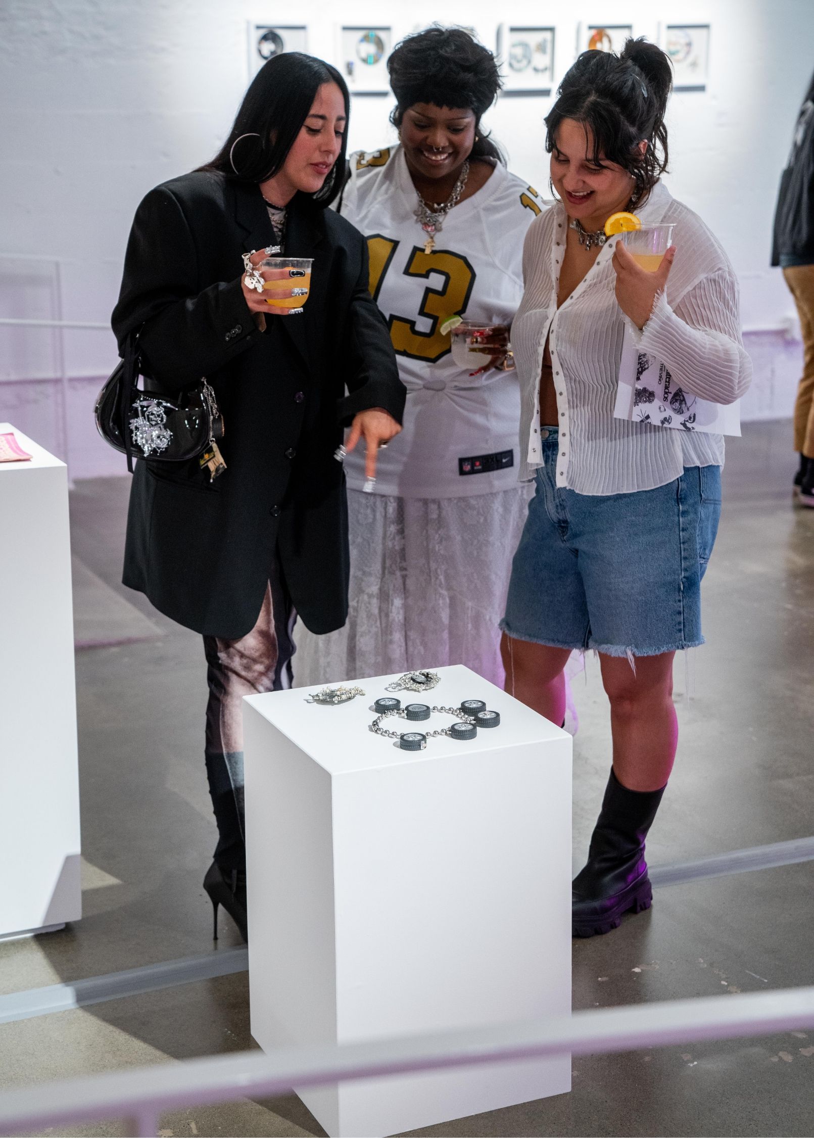 Three people stand around a white pedestal displaying objects in an art gallery. They are holding drinks and discussing the exhibit.