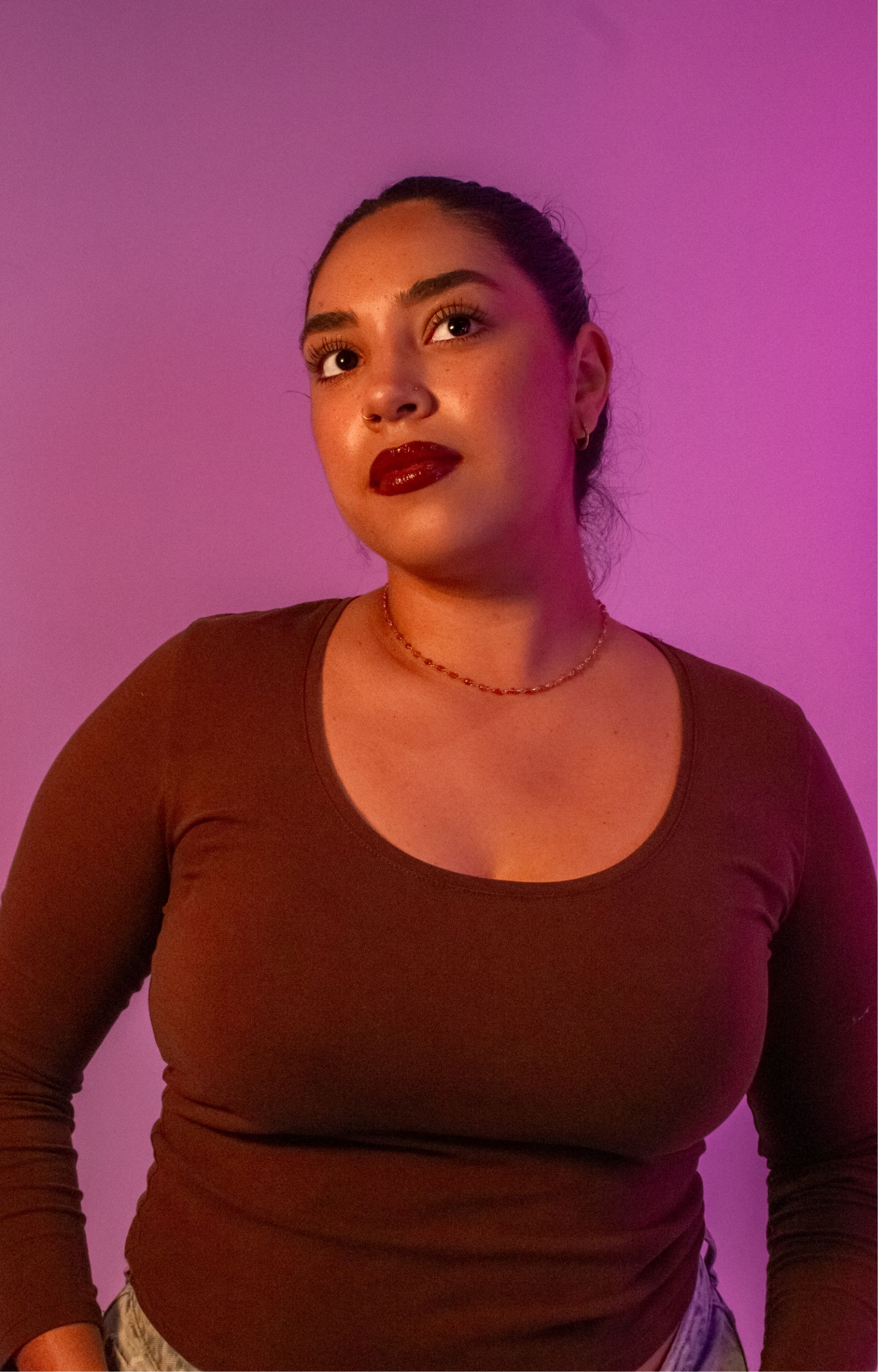 A woman wearing a brown top and a necklace stands against a purple-lit background, looking upwards with a focused expression.