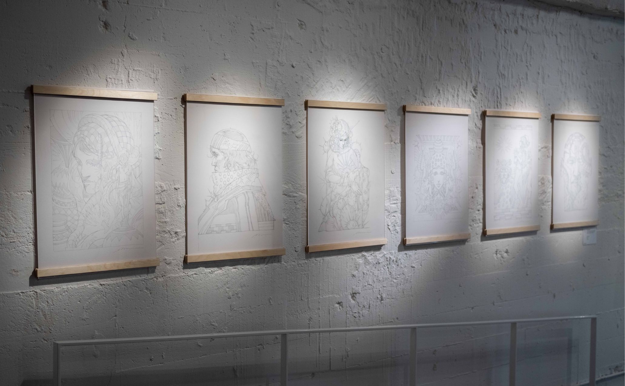 Six framed sketches are displayed on a white textured wall in an art gallery, each sketch subtly lit from above.