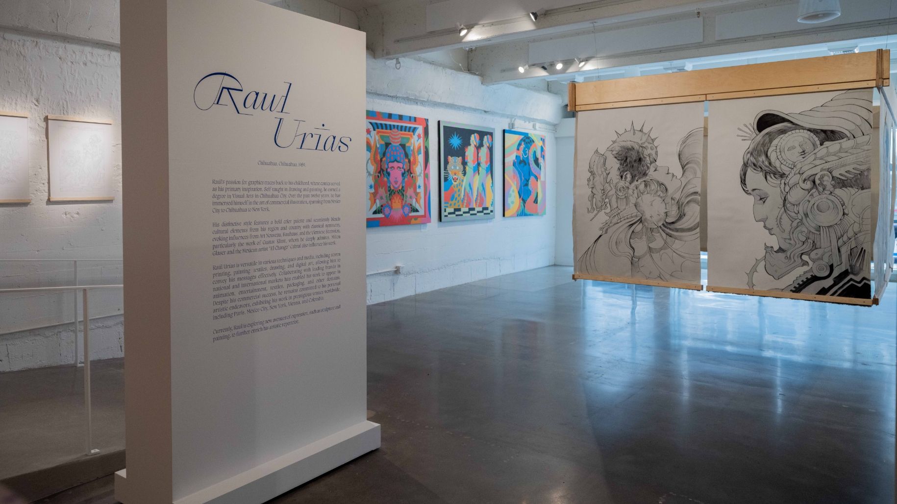 An art gallery display featuring a large sign with text about Raul Urias, colorful artworks on the walls, and intricate black-and-white drawings hung in the center.