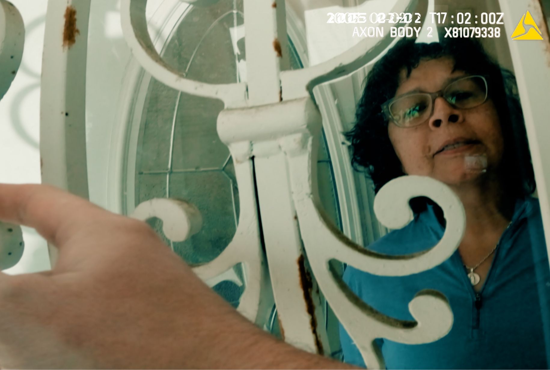 A person with curly hair and glasses stands behind a decorative metal door as a hand pushes it open. Timestamp and camera data overlay the image.