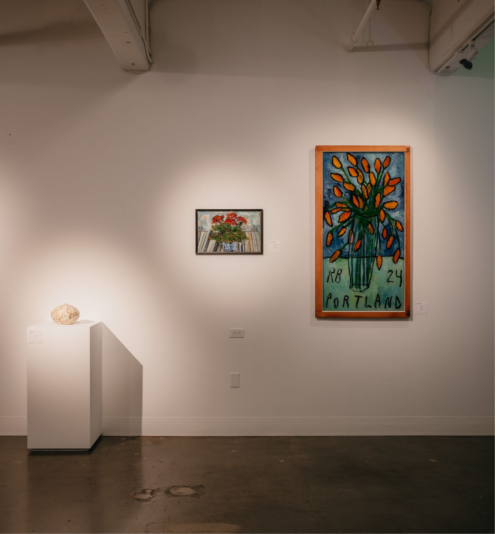 Gallery wall with three art pieces: a small framed painting of flowers, a medium painting with "PORTLAND" text, and a rock on a pedestal. Walls and floor are minimalist and well-lit.