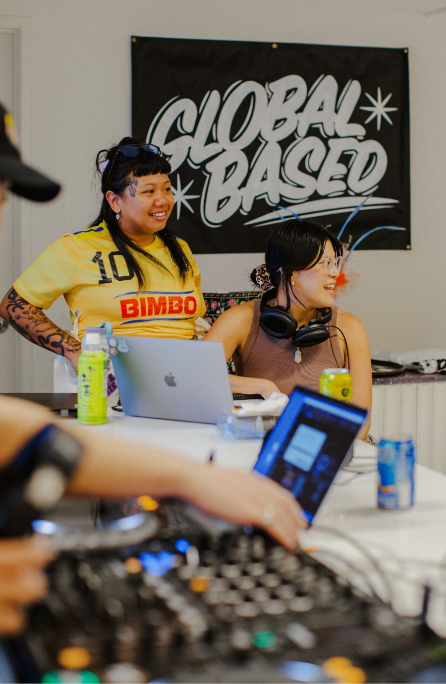 Two individuals sitting near DJ equipment and laptops in front of a "GLOBAL BASED" sign, with drinks and headphones on the table. One person is wearing a yellow top with "10 BIMBO" printed.