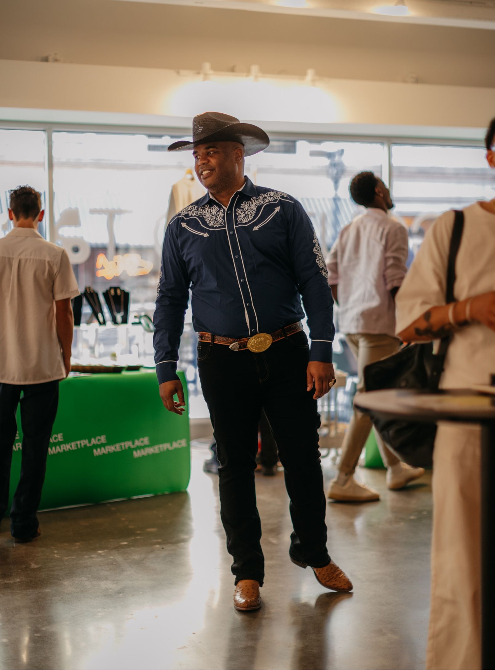 A man wearing a cowboy hat and a blue embroidered shirt stands inside a marketplace, surrounded by people and displays.