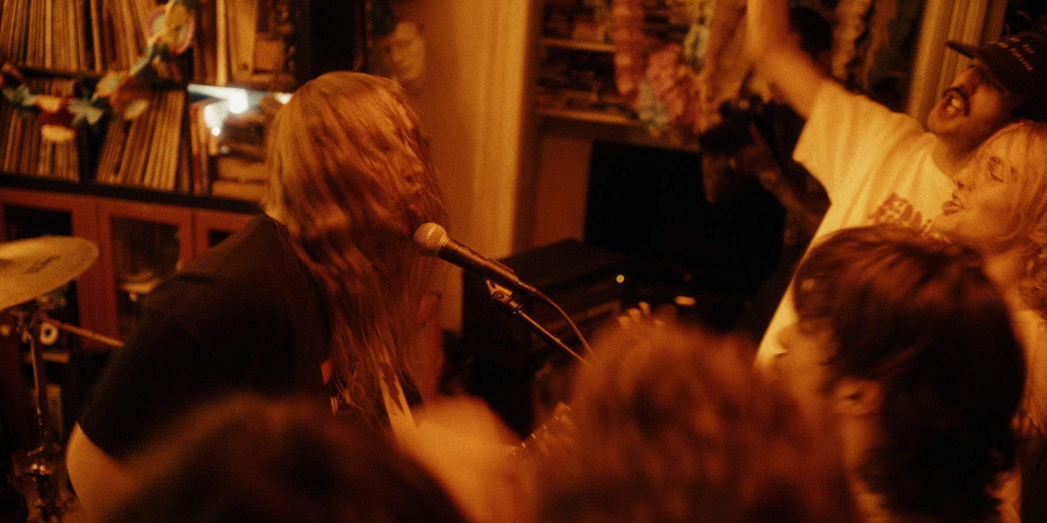 A band performs energetically in a dimly lit room filled with an enthusiastic audience. Records are visible in the background.